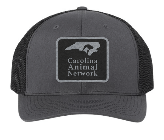 Charcoal/black trucker hat featuring the Carolina Animal Network logo in a black and silver leatherette patch. Front view.